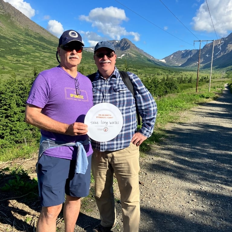 Two men hiking in Powerline Pass holding a sign in the shape of a plate that says